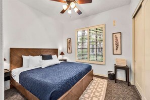 Un dormitorio con una cama grande, un ventilador de techo, una ventana con persianas y obras de arte enmarcadas en la pared.