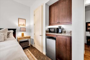 A compact hotel room with a bed, bedside lamp, a small table with a lamp, a microwave, and a refrigerator.