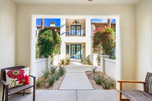 A pathway leading to a balcony with a ceiling fan, surrounded by greenery and a white wall.