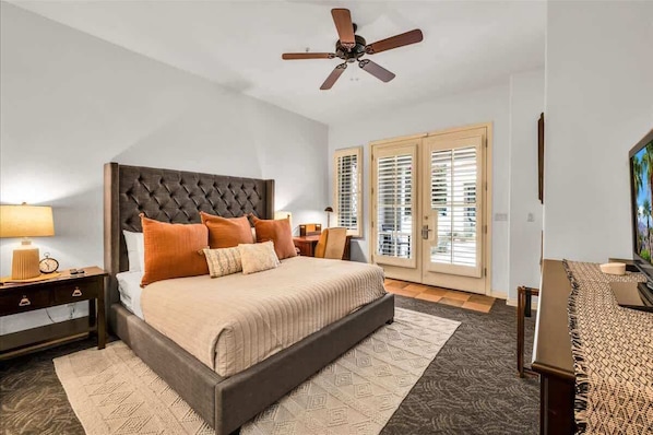 A bedroom with a tufted headboard, a bed with beige bedding, two nightstands, a desk with a chair, a ceiling fan, and a view through the glass doors.