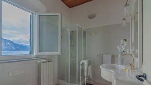 A bathroom with a glass-enclosed shower, a white sink with gold fixtures, and a view of the sea through a window. - Varenna Wonders  (Varenna)