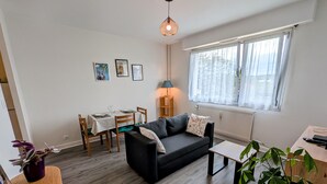 Una sala de estar moderna con un sofá negro, una mesa de comedor con sillas, una mesa de centro y un amplio ventanal con cortinas.