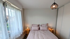 Un dormitorio con una cama, dos mesitas de noche con lámparas, una ventana con cortinas traslúcidas y un colgador con luz.