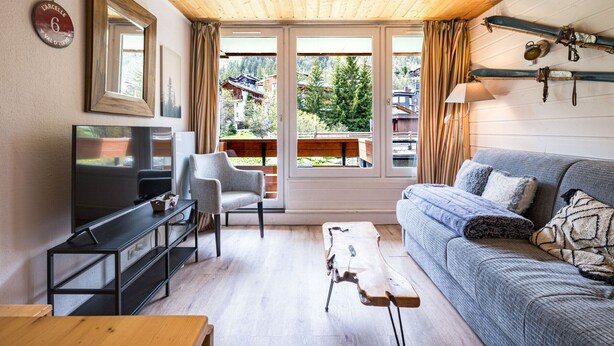 A cozy living room with a wooden ceiling, a sofa, a TV, a chair, and a window with a view of buildings and trees.