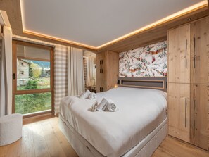 A bedroom with a large bed, wooden wardrobe, and a window with a view of buildings and trees.