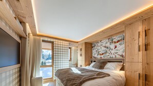 Ein modernes Schlafzimmer mit einem großen Bett, Holzbalkendecke, einem Fenster mit Aussicht und einem an der Wand befestigten Fernseher.
