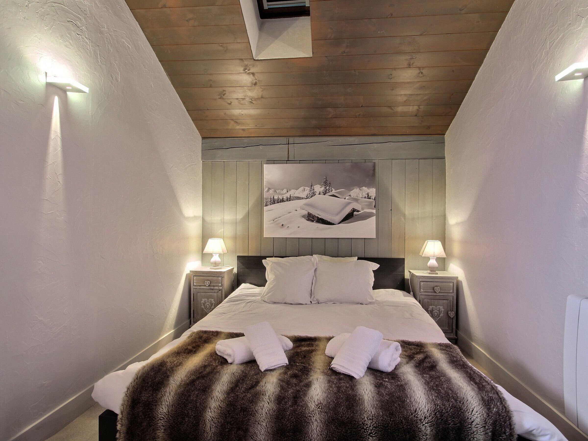 Un dormitorio con techo de madera, una cama con ropa blanca, dos lámparas de noche y un cuadro enmarcado en la pared.