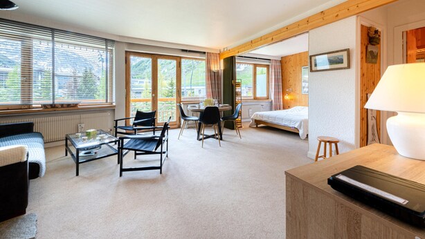 A well-lit room with a bed, a desk, and a chair. There are large windows providing natural light and a view of the outdoors.