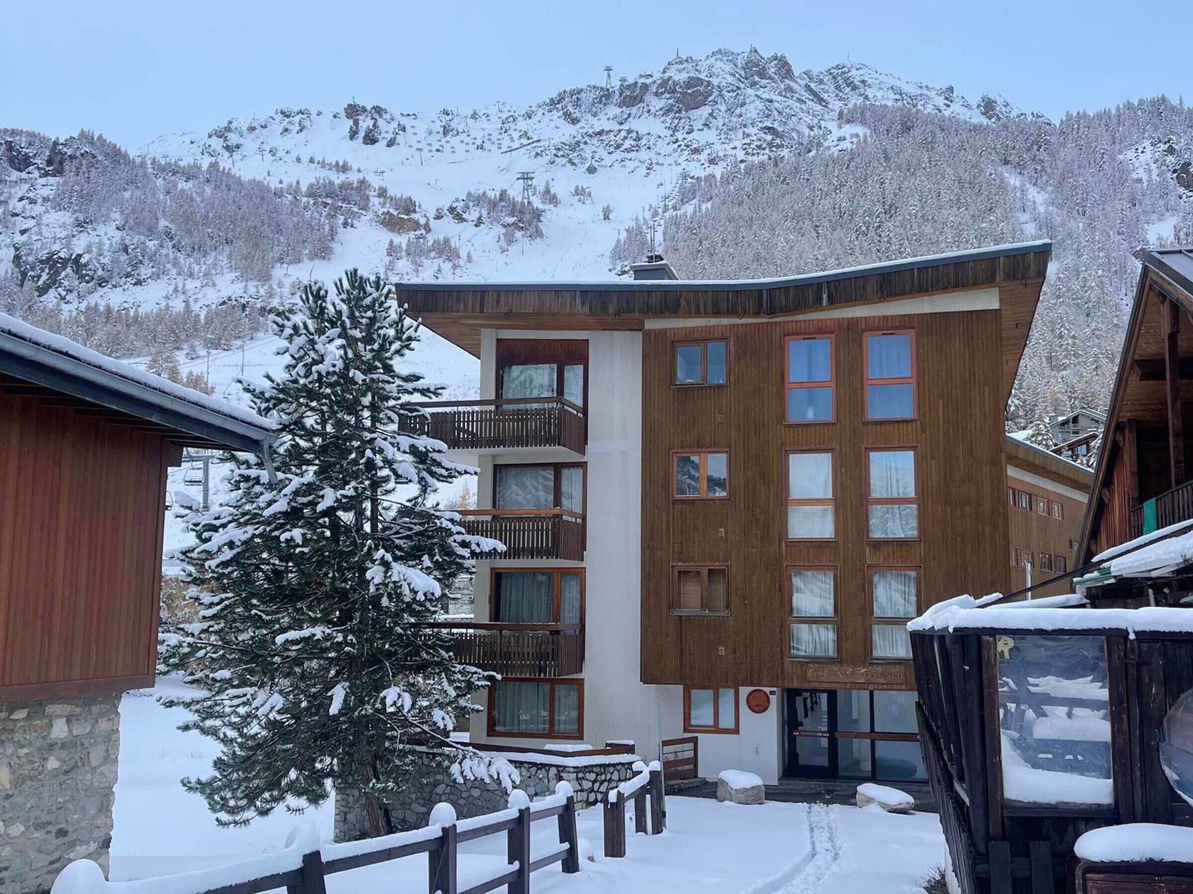Un bâtiment en bois à plusieurs étages, doté de balcons, d’un mur en pierre et surplombant un paysage enneigé avec des montagnes en arrière-plan.