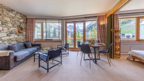 A cozy room with a stone wall, a sofa, a dining table with chairs, and large windows offering a mountain view.