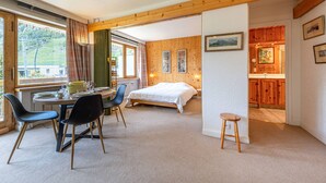 A room with a bed, a dining table with chairs, a small stool, and a kitchen area with wooden cabinets.