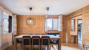 Ein Zimmer mit Holzwänden, einem großen Esstisch, Stühlen, Pendelleuchten und einem Ausblick durch ein Fenster.