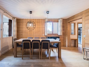 A wooden-paneled room with a large dining table, chairs, pendant lights, and a view through a window.
