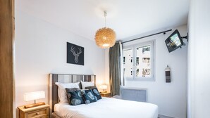 Ein Schlafzimmer mit Bett, Nachttischen, einem an der Wand befestigten Fernseher, einer Deckenleuchte und einem Fenster mit Vorhängen.