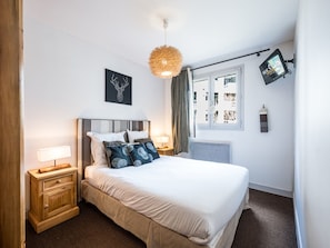 A bedroom with a bed, bedside tables, a wall-mounted TV, a ceiling light fixture, and a window with curtains.