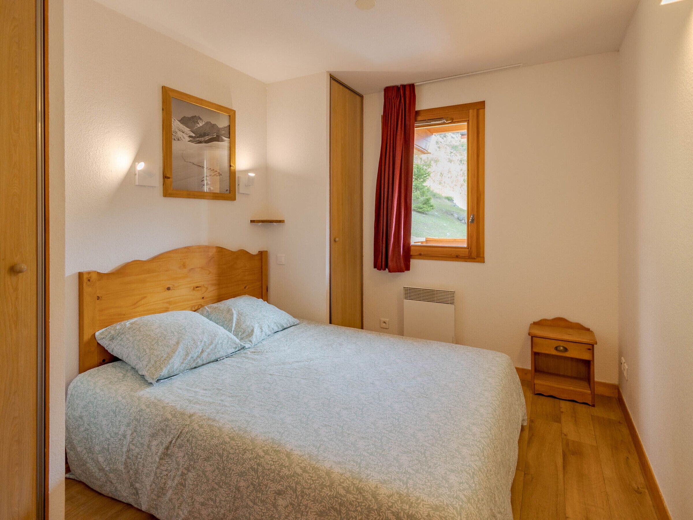 Ein Schlafzimmer mit einem hölzernen Bett, einem kleinen Nachttisch, einem Fenster mit rotem Vorhang und einem Wandheizkörper.