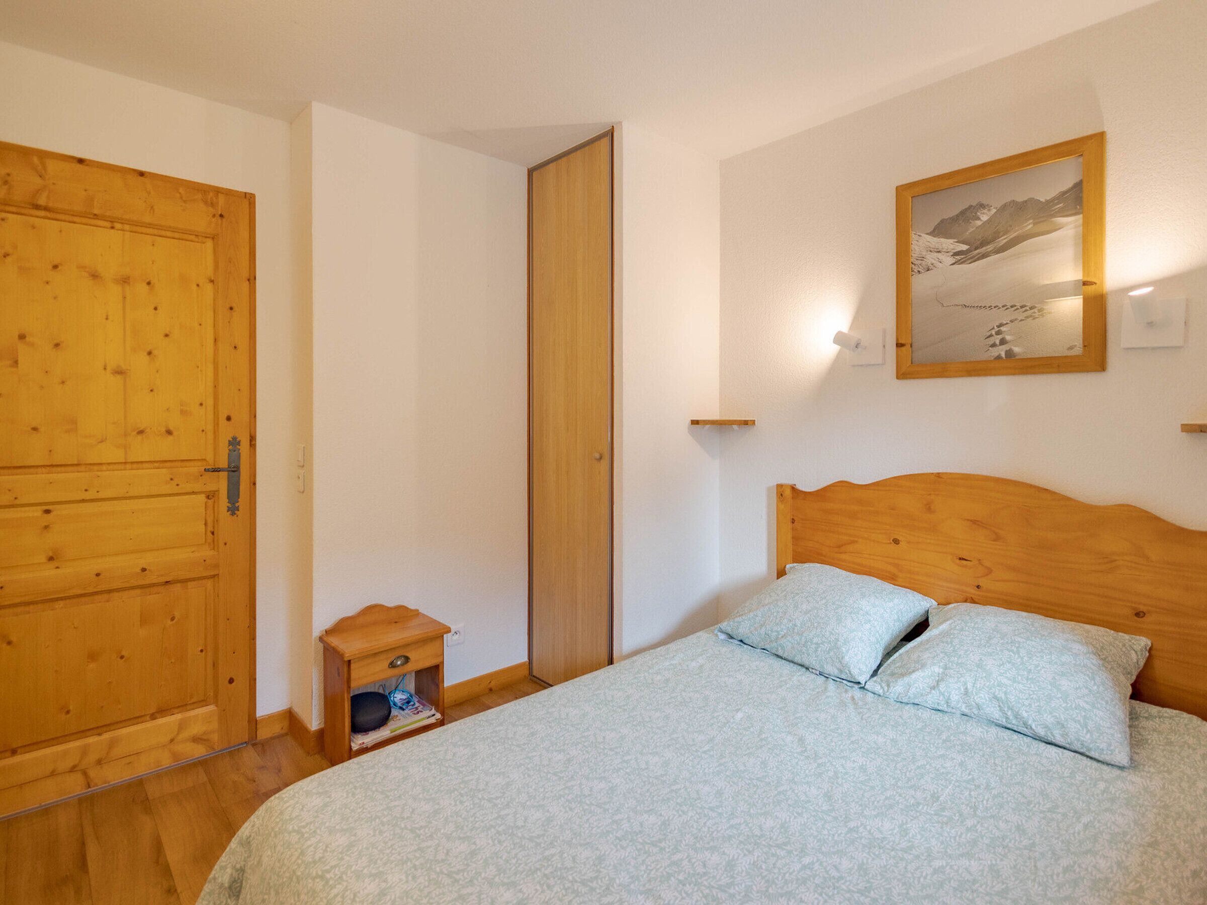 Ein Schlafzimmer mit Holztür, Nachttisch mit Lampe, Bett mit zwei Kissen und einem gerahmten Bild an der Wand.