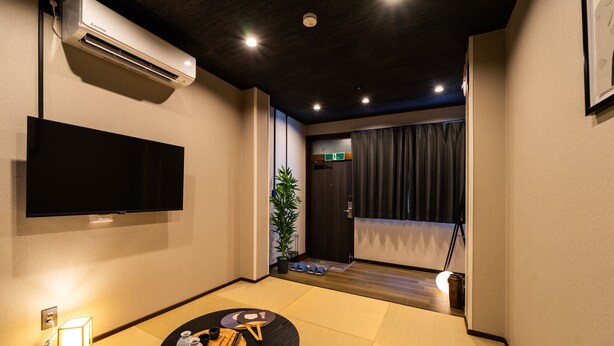 Una habitación de hotel moderna con televisión de pantalla plana, una pequeña mesa de comedor con palillos y una vela, suelo de tatami y una puerta corredera que da a un balcón.