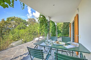 A covered outdoor patio with a stone floor, green metal chairs, a table set for dining, and a view of trees and a building in the background. - Honeymoon Love Retreat 2 (Minusio)