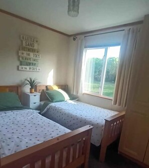 Un dormitorio con dos camas individuales, una mesita de noche con lámpara, un cartel en la pared y una ventana con cortinas.
