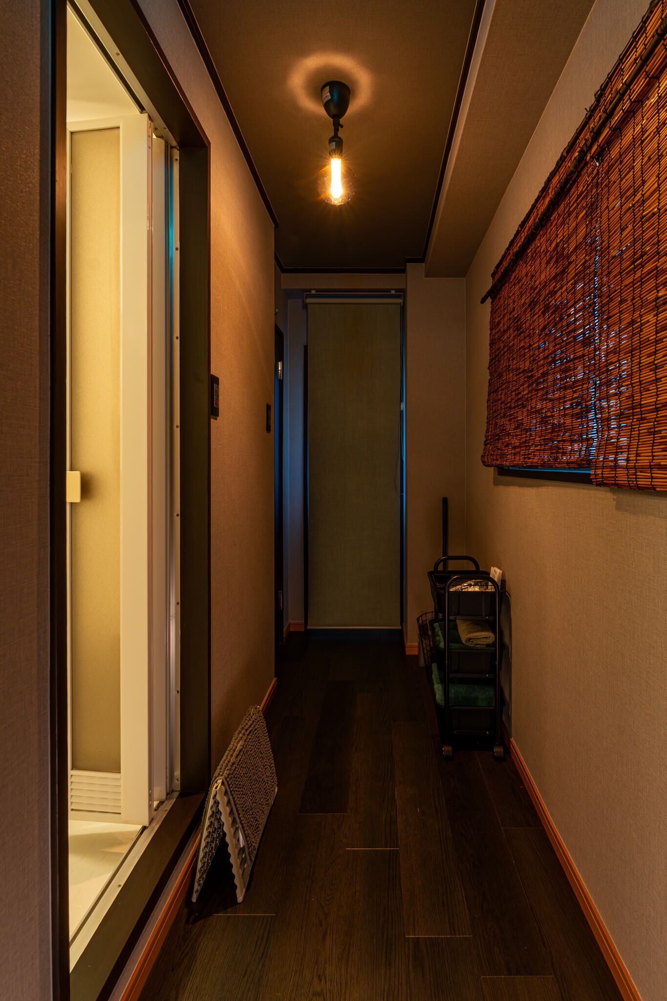 A narrow hallway with a wooden floor, a ceiling light, a door at the end, and a basket with items on the right side.