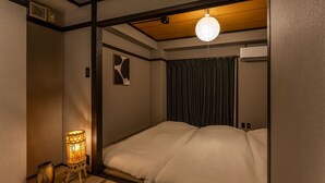 A traditional Japanese-style room with tatami flooring, a shoji screen, a futon bed, a decorative lamp, and a small table with a vase. - Oyado Kinokuniya Shindaikumachi (Nagasaki)