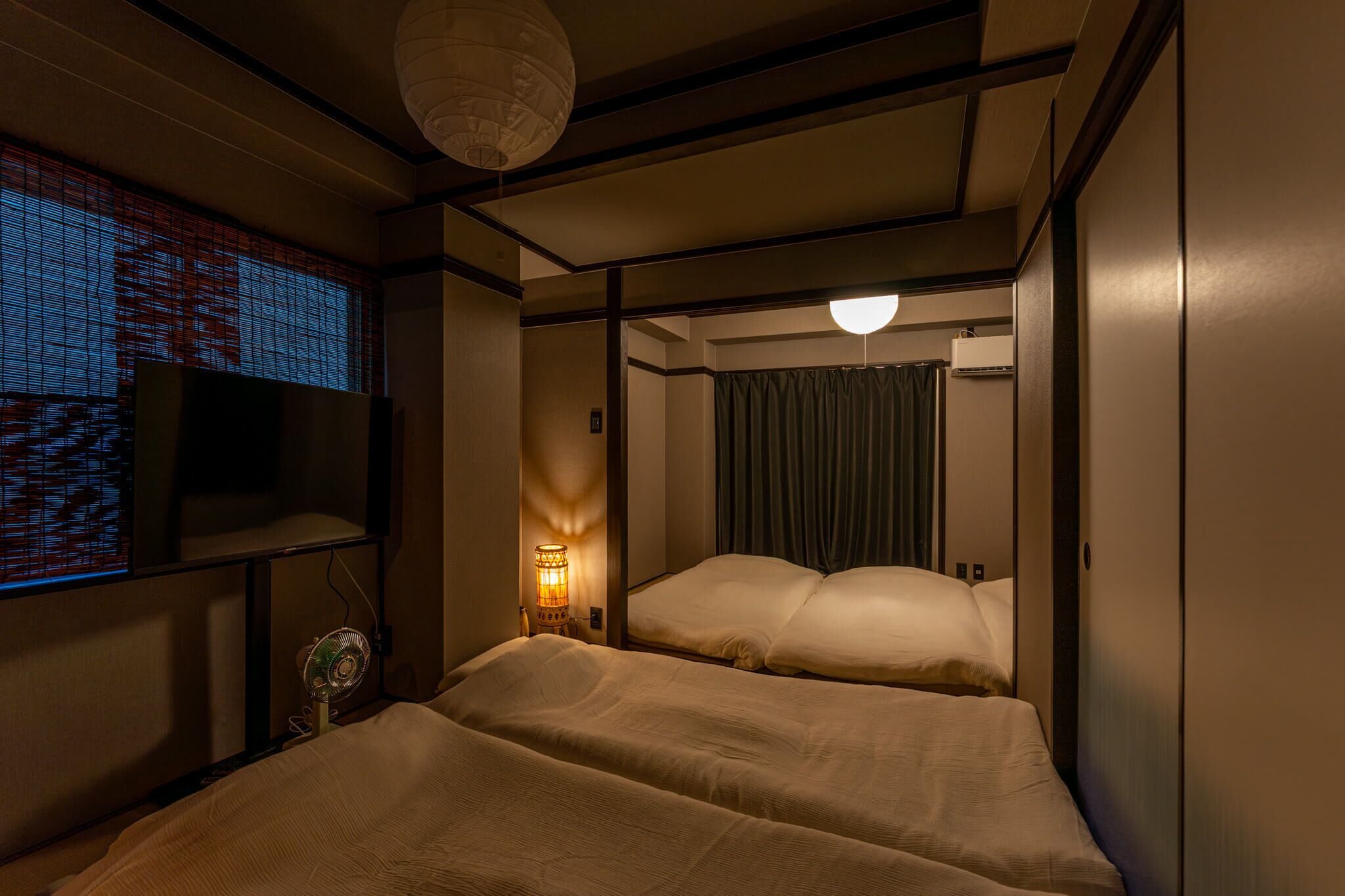 Habitación de hotel con dos camas, un televisor en la pared izquierda, un ventilador de pie y una lámpara del techo.