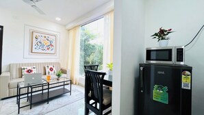 Een moderne woonkamer met een bank, een glazen salontafel, een laptop en een eethoek met stoelen en een tafel. Er staat een zwarte koelkast met een sticker erop, en een raam met gordijnen.