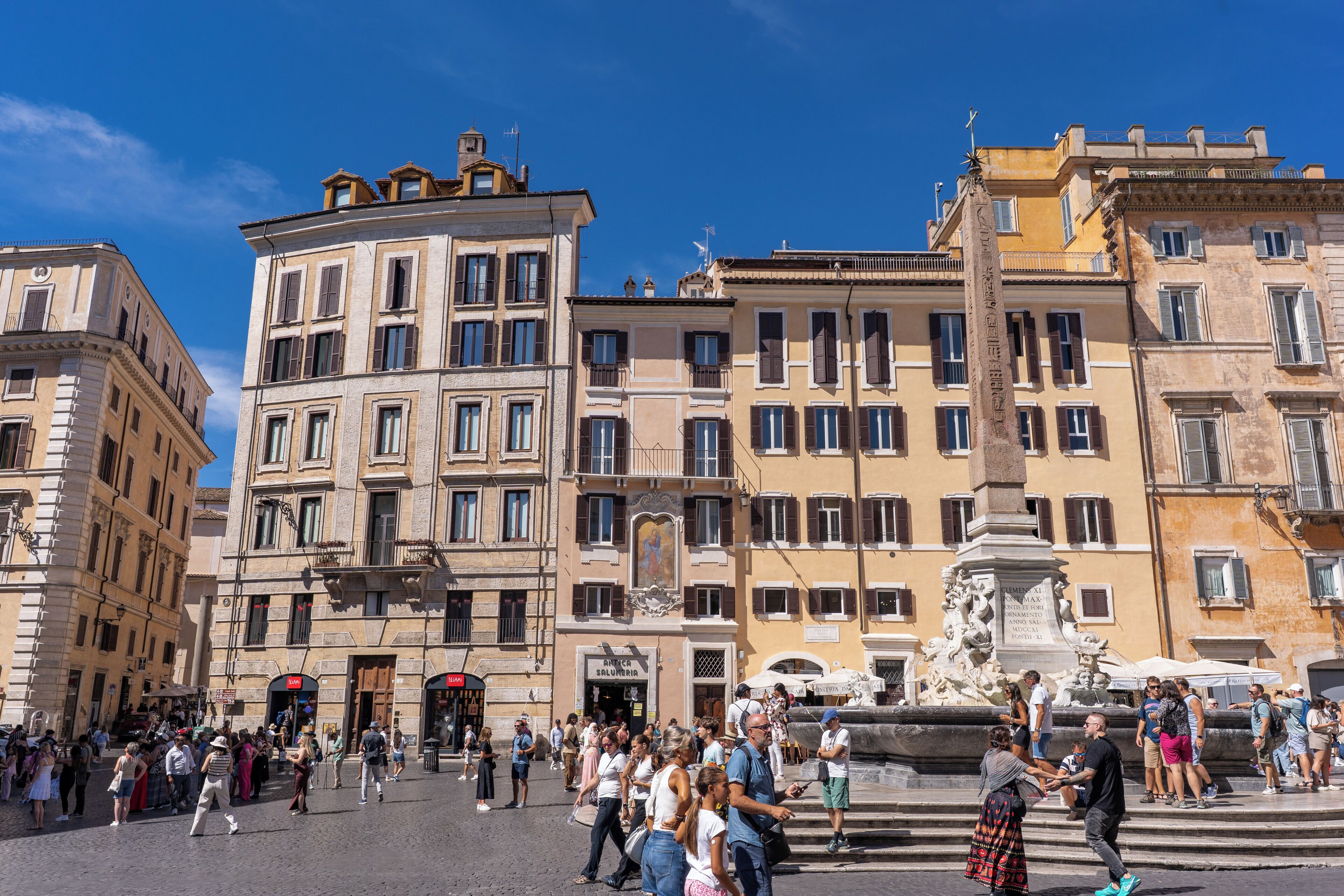 A historic square with classical architecture, a fountain, and a crowd of people.