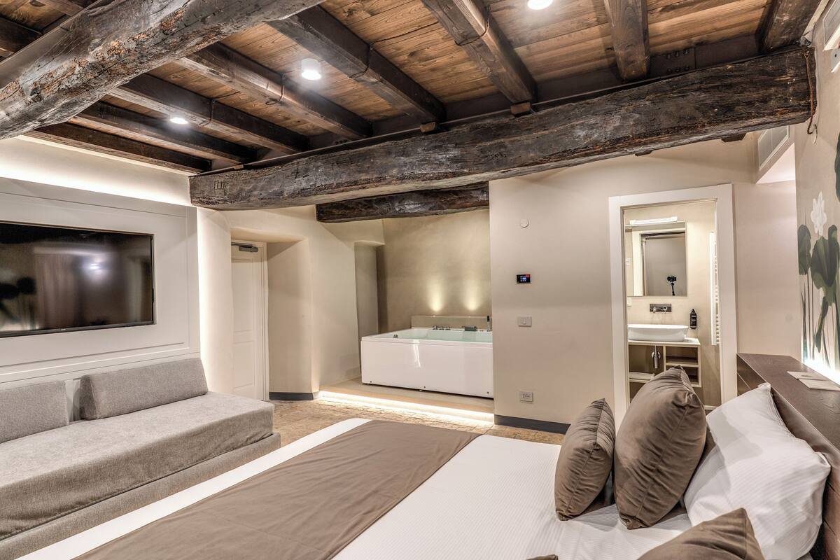 A bedroom with a large bed, a sofa, a TV, and a bathroom visible through an open door.