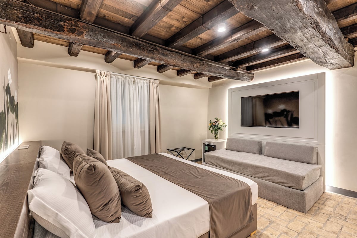 A hotel room with a wooden ceiling, a bed with brown bedding, a sofa, a small table with a vase, and a painting on the wall.