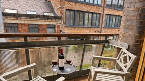 A wooden deck with two white chairs and a table, a bottle of wine, and a view of a brick building with windows. - Apt 4,34 Wood street (Liverpool)