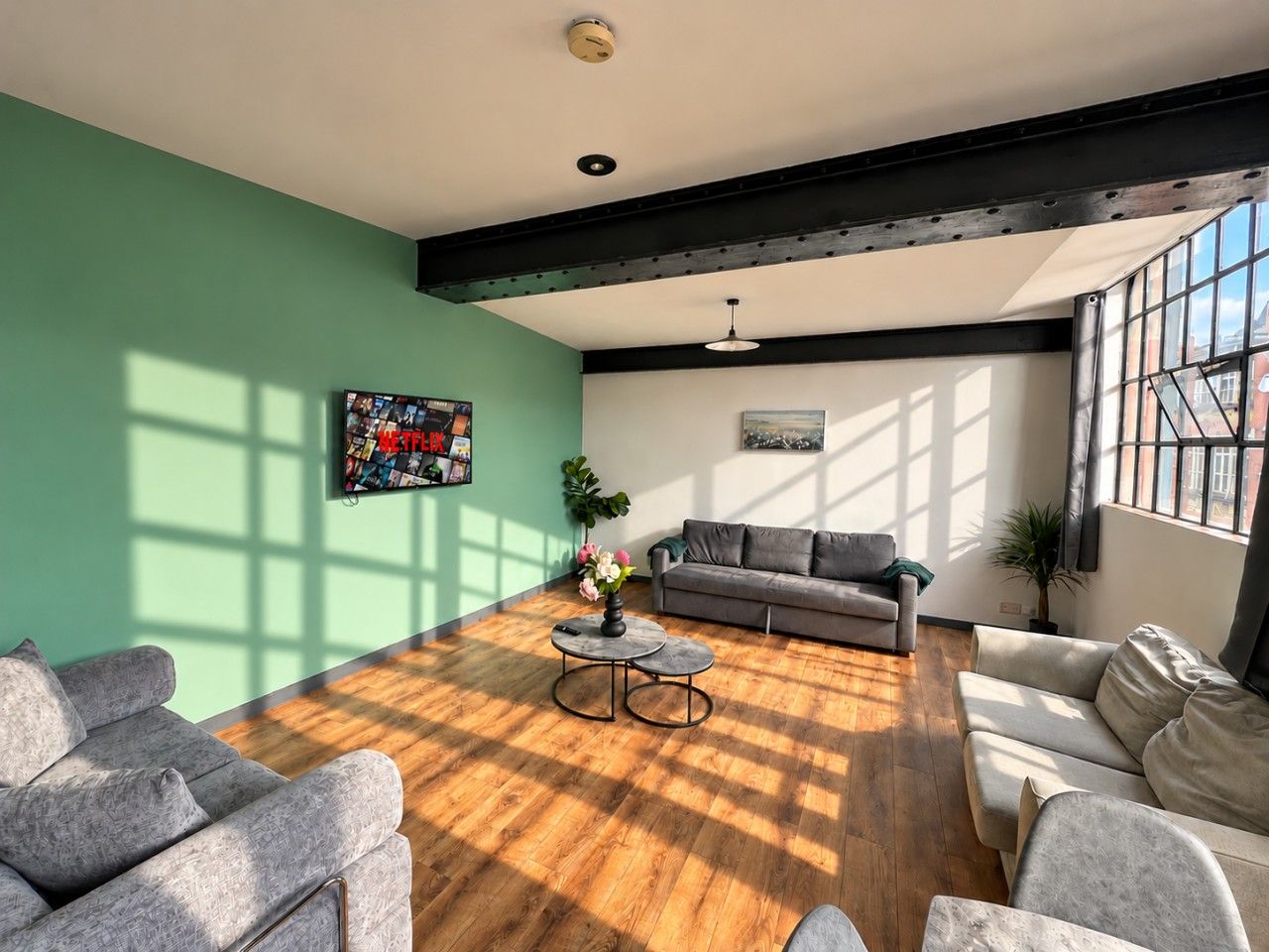 Ein geräumiges Wohnzimmer mit Holzboden, einem grauen Sofa, einem Couchtisch und einem großen Fenster, das für natürliches Licht sorgt.