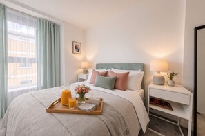 Habitación de hotel con cama, mesitas de noche, bandeja con jugo de naranja, florero con flores y ventana con cortinas.