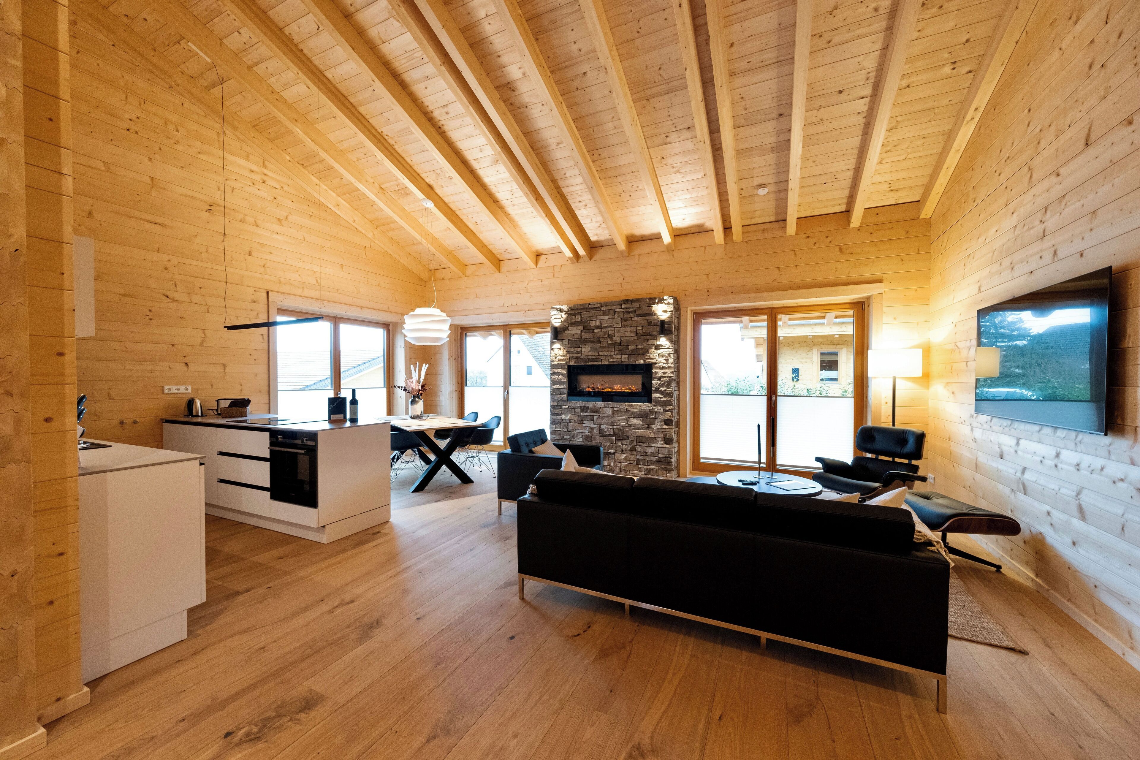 A spacious living area with a fireplace, wooden ceiling beams, a modern kitchen, and a large TV mounted on the wall.