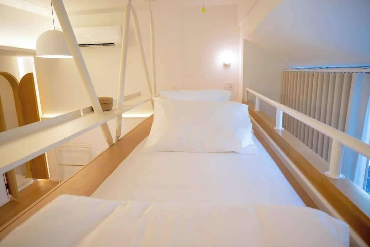A hotel room with a wooden floor, white bedding, a white railing, and a window with blinds.