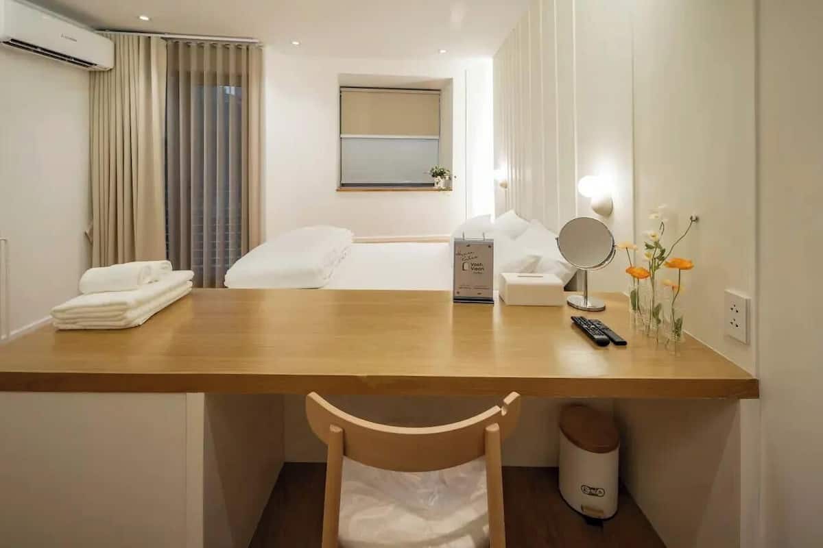 A hotel room with a wooden desk, a bed with white linens, a bedside table with a lamp and flowers, a window with curtains, and an air conditioning unit.