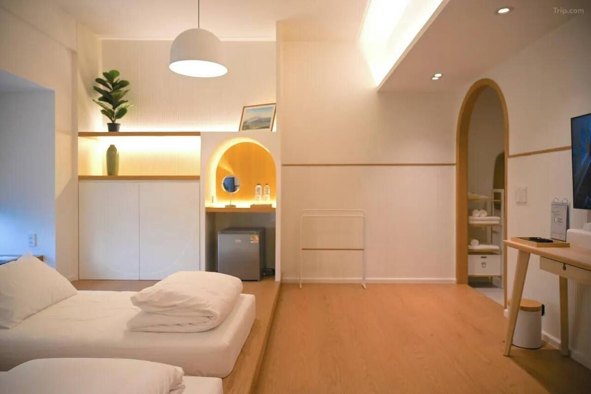 A hotel room with a bed, bedside table, lamp, and a small desk with a chair. There is a plant on the bedside table and a shelf with a plant and a picture frame.