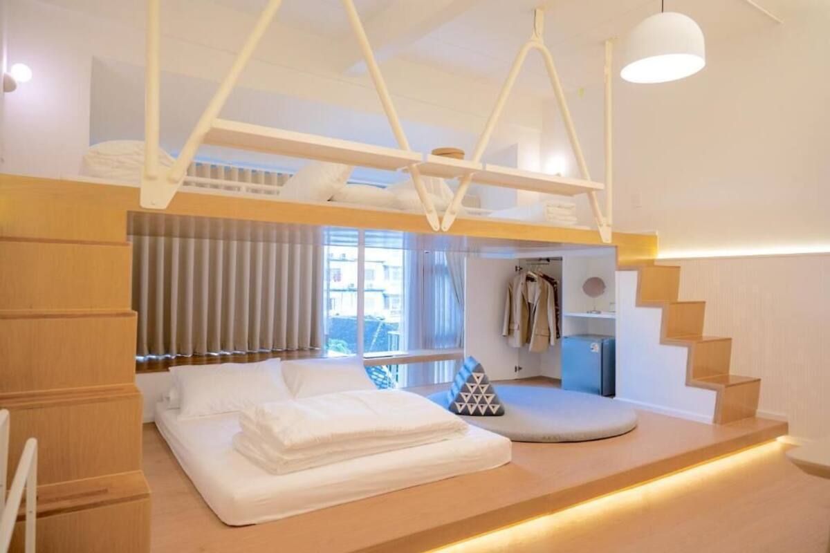A modern hotel room with a bunk bed, a seating area with cushions, a staircase, and a wardrobe.