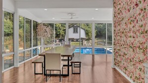 Amplia habitación con una gran mesa de comedor y bancos, ventanas amplias que ofrecen vistas a una piscina y una casa.