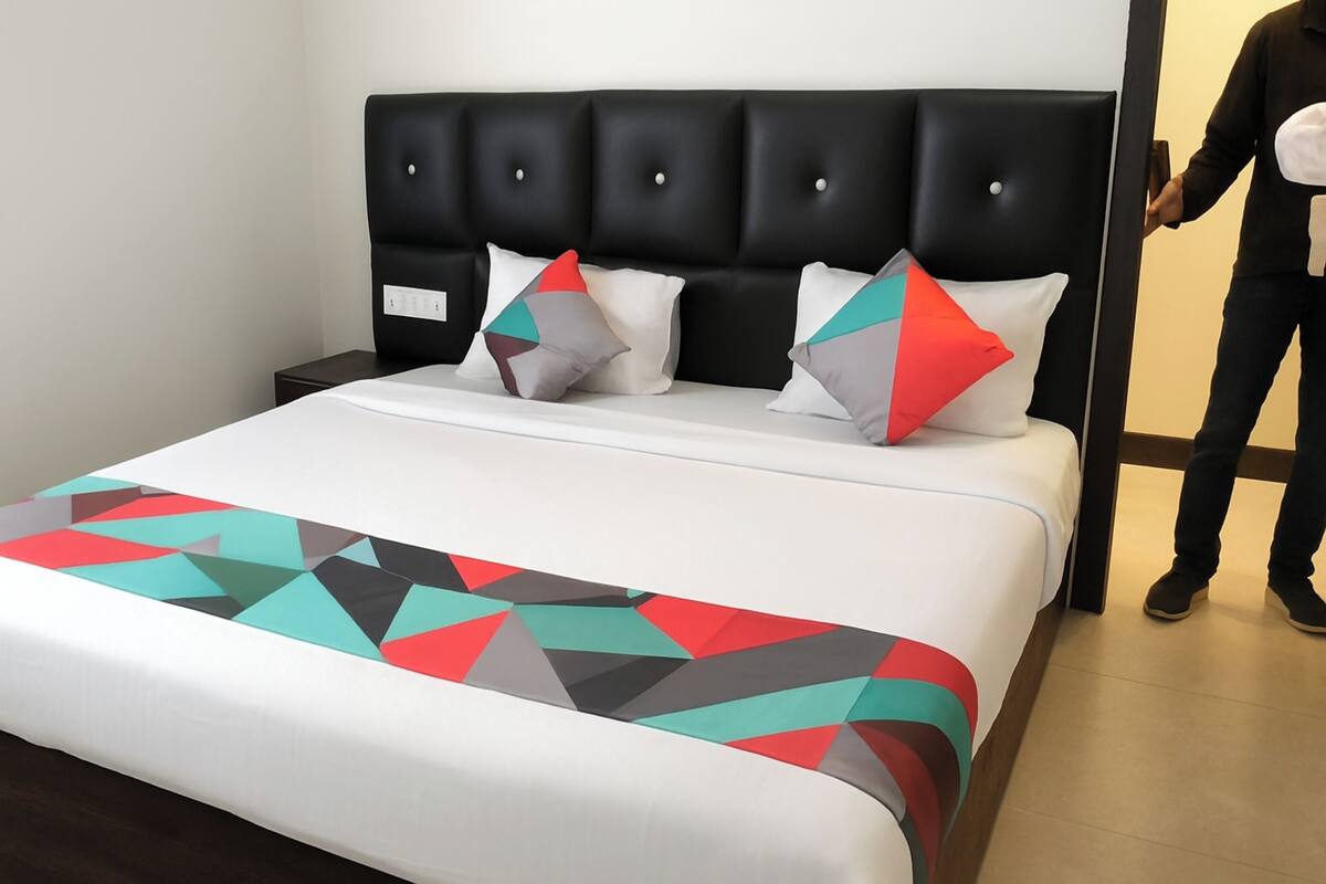 A hotel room with a bed, a black upholstered headboard, and a white bedspread with a colorful geometric pattern.