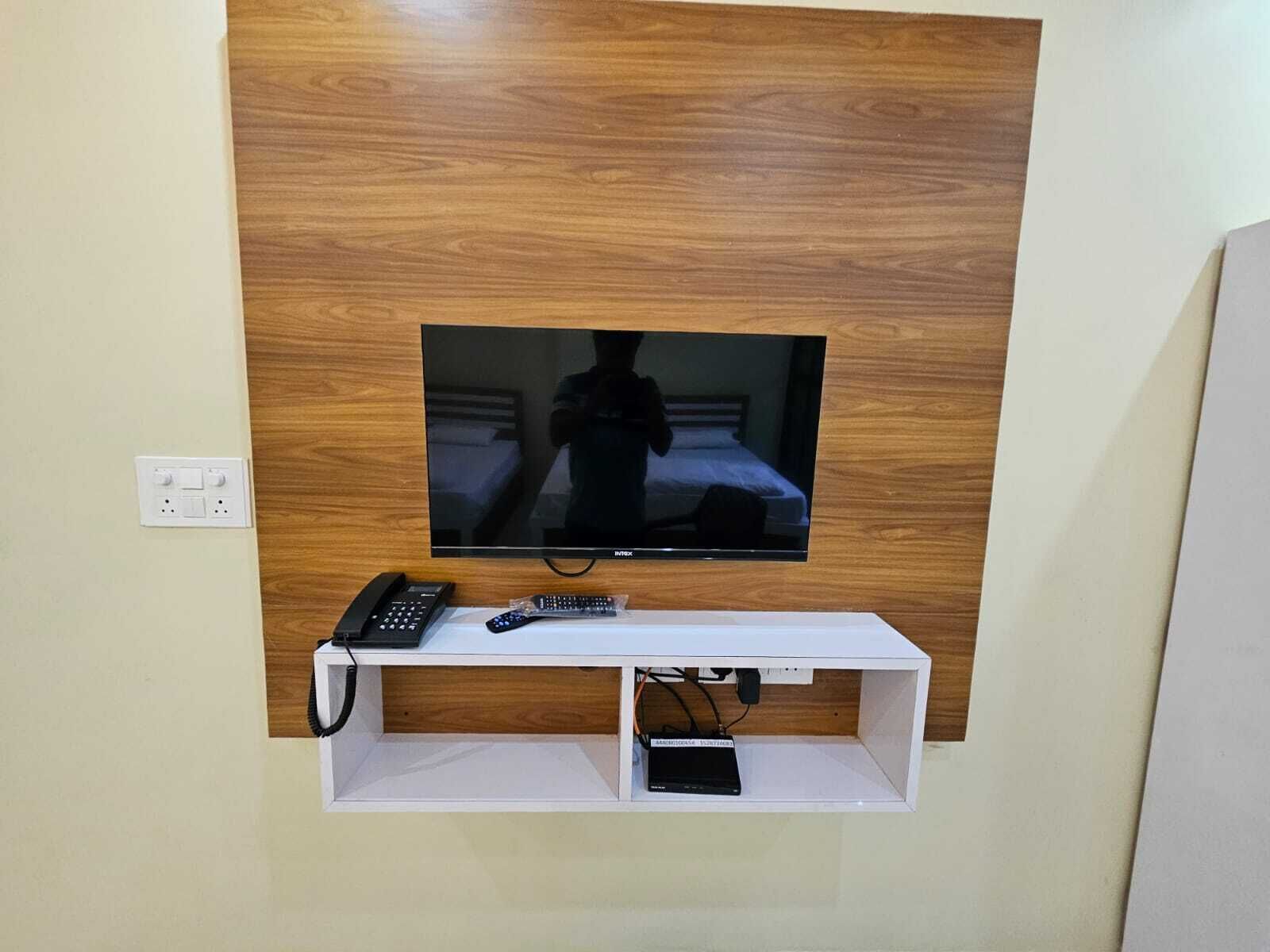 A television mounted on a wooden wall with a white shelf below containing a telephone, remote controls, and a power strip.