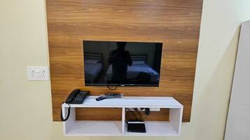 A television mounted on a wooden wall with a white shelf below containing a telephone, remote controls, and a power strip.