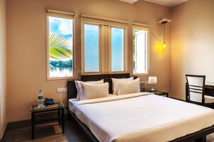 Habitación de hotel con una cama grande, dos mesitas de noche, una silla y una ventana con vistas a palmeras.