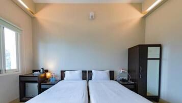 Quarto de hotel com duas camas, uma escrivaninha com luminária, um guarda-roupa e uma janela.