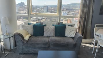 A living room with a grey sofa, a black coffee table, and a view of a cityscape through large windows.
