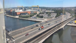 A multi-level highway with several lanes of traffic, a bridge crossing over a body of water, and a large stadium with a distinctive orange and white color scheme. - City Centre high rise (Belfast)