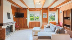 Een woonkamer met houten balken, een open haard, een televisie, een bank en een raam met uitzicht op groen.