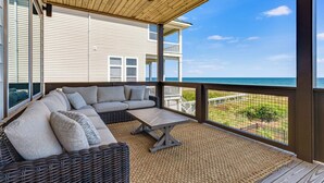 Un espace extérieur aménagé, abrité et doté d'un canapé en osier et d'une petite table, offrant une vue sur une plage et une maison avec balcon.
