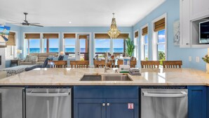 Une cuisine dotée d’un grand îlot central, un coin repas offrant une vue sur l’océan et un salon avec un ventilateur de plafond.